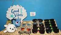 Full lot displayed including multiple glasses and sunglasses in black, brown, and green frames, two plush toys (white round seal and blue fish), and a blue and white wooden hanging sign reading 'Good Vibes Only' with starfish decoration.