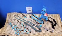 Set of necklaces arranged on woven mat with blue background, along with a blue TY Beanie Baby Stuffy plush toy.