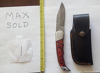 Photo showing the Damascus folding knife with wood handle placed next to a measuring tape and a black leather sheath. The blade shows characteristic Damascus patterning. The handle is reddish wood with metal bolsters. The sheath has stitching and a snap button.