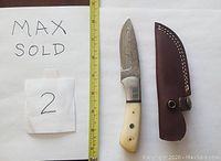 Damascus straight knife with camel bone handle and accompanying brown leather sheath next to a measuring tape for scale, showing the blade with visible Damascus pattern and camel bone handle with rivets.