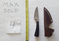 Damascus steel straight knife with buffalo horn handle placed next to a measuring tape, accompanied by a brown leather sheath. The knife blade shows layered Damascus pattern and the handle has brass rivets.