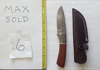 Photo shows Damascus straight knife with wood handle featuring mosaic pins and brown leather sheath. A yellow measuring tape alongside indicates blade length.