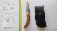 Photo showing a Damascus folding knife with wood handle next to a leather sheath. A tape measure adjacent shows knife length about 8 inches open. Included is a paper with 'Max Sold 8' written.