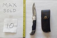 Damascus folding knife with buffalo horn handle displayed next to a measuring tape and its leather sheath to the right. The blade shows the characteristic Damascus pattern. The sheath is black with stitching and a button clasp.