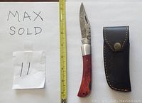 Photo of the Damascus folding knife alongside a black leather sheath, displayed next to a yellow measuring tape for scale measurement, showing details of the blade pattern and wood handle.