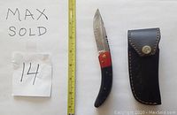 Damascus folding knife with buffalo horn and wood handle positioned next to a measuring tape and next to a black leather sheath with brass button.