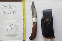 Photo showing the Damascus folding knife and leather sheath alongside a measuring tape for scale and a paper with 'MAX SOLD 15' written on it.