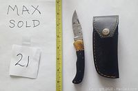 Image showing Damascus folding knife with buffalo horn handle and brass bolster next to a closed black leather sheath. A measuring tape and handwritten note indicating MaxSold and lot number 21 are also visible.