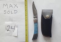 Knife with patterned Damascus steel blade, dyed blue bone handle, and black leather sheath, against a white background with measuring tape.