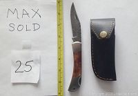 Photo showing Damascus folding knife with died bone handle next to black leather sheath, measuring tape for scale, and paper labeled Max Sold 25.