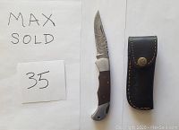 Photo of a Damascus folding knife with an exposed patterned blade, wood handle, and metal bolsters, placed next to a black leather sheath and a paper with 'MAX SOLD 35' written.
