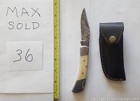 Photo showing the Damascus folding knife with a camel bone handle and a black leather sheath next to a ruler for size reference.