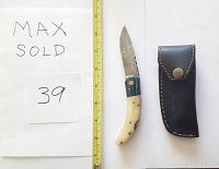 Damascus folding knife with ivory-colored camel bone and dark wood handle placed next to a yellow measuring tape and black leather sheath with stitched edges and snap button.