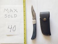 Damascus folding knife with dyed blue bone handle beside a measuring tape and leather sheath closure shown.