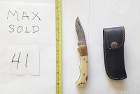 Damascus folding knife with camel bone handle next to yellow measuring tape and black leather sheath.