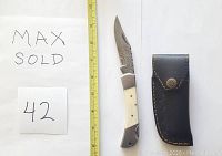 Damascus folding knife alongside a black leather sheath, placed next to a ruler for size reference.