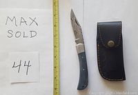 Damascus folding knife with dyed blue bone handle next to measuring tape, and black leather sheath.