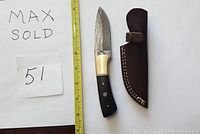 Top-down photo showing the straight knife with visible Damascus blade pattern, buffalo horn handle with mosaic pins and the brown leather sheath next to it. A measuring tape and a handwritten MaxSold 51 sign included.