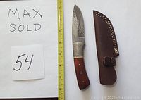 Full view of the Damascus straight knife with wood handle and attached sheath beside a measuring tape showing size approximately 8.5 inches total length.