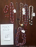 Variety of pearl-style necklaces in shades of red, pink, white, blue, purple, and multicolored beads laid flat on a surface with visible price tags. Includes a sterling black dog tag on a black chain and a silver-tone ring.