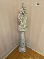 Full view of cast statue seated atop the tall decorative pedestal stand in corner of room.