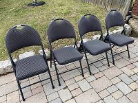 Four black fabric padded folding chairs set in a row outdoors on a stone paved surface with grass and stone edging visible behind them.