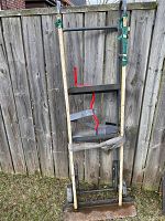 Full view of appliance dolly standing upright against fence outdoors, showing white handles, frame and straps.