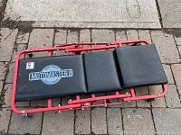 Top view of the Motomaster creeper dolly showing the red metal frame and three black vinyl or leather cushioned pads. The largest pad displays the Motomaster logo.