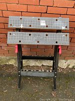 Front view of folding work bench closed against a brick wall, showing two rectangular top panels with measurement markings and holes.