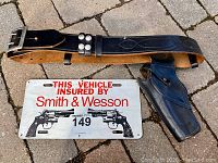 Image showing full length of black leather gun belt with silver buckle and rivets, with metal license plate below it.