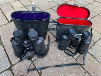 Two pairs of binoculars with their respective open cases on a paved surface, showing the exterior and interior velvet lining of the cases.