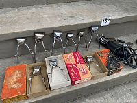 Seven metal hand operated hair clippers displayed in a row with four original boxes underneath. An electric hair clipper is shown at the right edge.