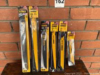Photo showing six DeWalt reciprocating saw blades in original packaging standing against a brick wall background on a wooden surface.