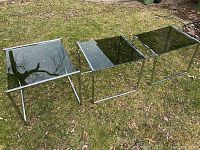 Set of three modernist chrome and glass tables arranged side by side outside on grass showing top and frame design.