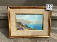 Full view of framed oil painting showing coastal cliffs and ocean with beige fabric matting and gold frame.