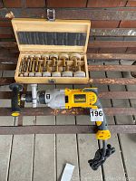 DeWalt hammer drill with yellow and silver body and black side handle alongside wooden case of drill bits.