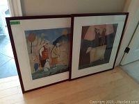 Two framed and matted prints leaning on the floor near a doorway, showing the overall lot display and frame style.