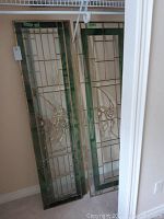 Two stained glass panels leaning against a wall, showing the hummingbird design and brass framing with green and clear glass sections.