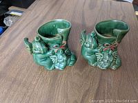 Two green pottery planters shaped with elephant figures hugging each vase, glossy finish with red bow detail