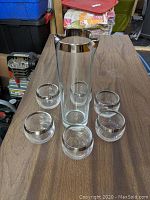 Clear glass double spout cocktail pitcher with silver colored rim and six matching old fashioned roly poly tumblers with silver colored rims on wood surface.