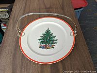 Top view of the round ceramic plate showing a Christmas tree with gifts and a red border, featuring the removable metal handle attached.