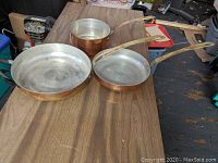 Set of three copper pans on wooden surface: two skillets and one saucepan, showing good overall condition without lids.