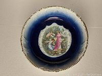 Full top-view photo of the porcelain bowl showing detailed neoclassical scene of two women in Grecian gowns with cherub, deep cobalt blue flow blue glaze fading to white at edges, scalloped and embossed decorative rim.