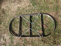 Top-down view of oval metal pot rack with two crossbars and hanging chains, placed on grass outdoors.