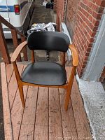 Front view of wooden armchair with black vinyl seat and backrest, showing curved armrests and tapered legs on wooden deck.
