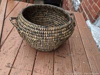 Round coiled grass basket with layered woven pattern and side handles shown on a wooden surface.