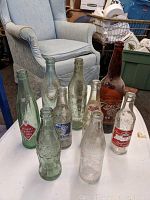 Photo showing 9 vintage soda bottles on white table with armchair and household items in background.
