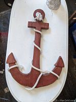 Carved wooden anchor wall hanging laying on white surface, showing front with white rope wrapped around the anchor.