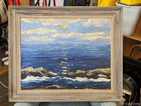 Framed oil painting showing a seascape with waves, rocks, a cloudy blue sky, and a sun reflecting on the water surface.