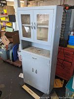 Front view of a tall steel cabinet hutch with glass doors on the top section, central drawer, and solid double doors below; showing wear and paint chipping.